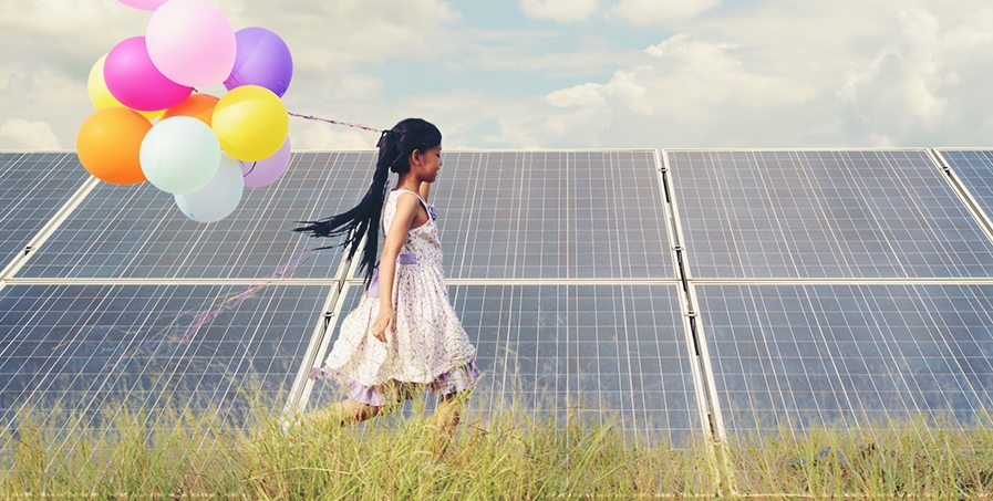 Young girl with dark hair and white sundress walking with a bunch of colorful balloons by a wall of solar panels in a grassy field.
